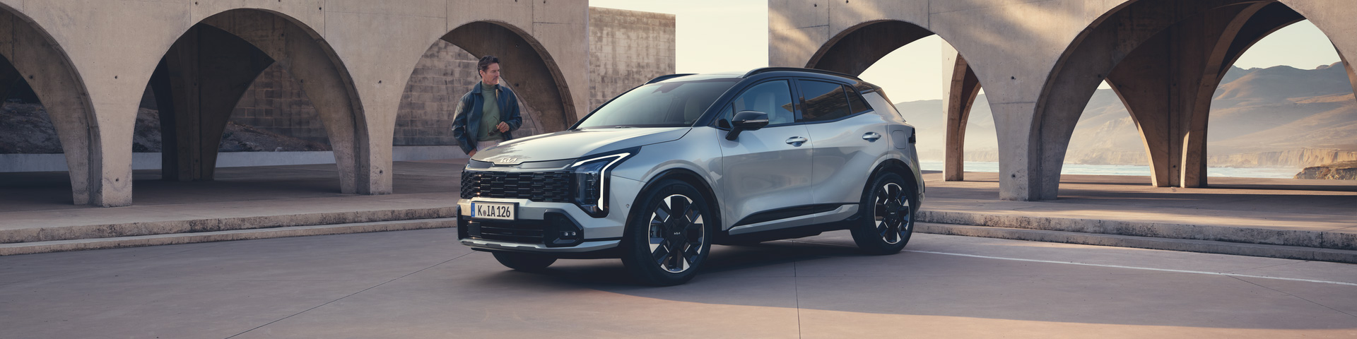 Der Kia Sportage HEV GT-line MY26 vor moderner Architektur mit einem männlichen Talent zur Seite.